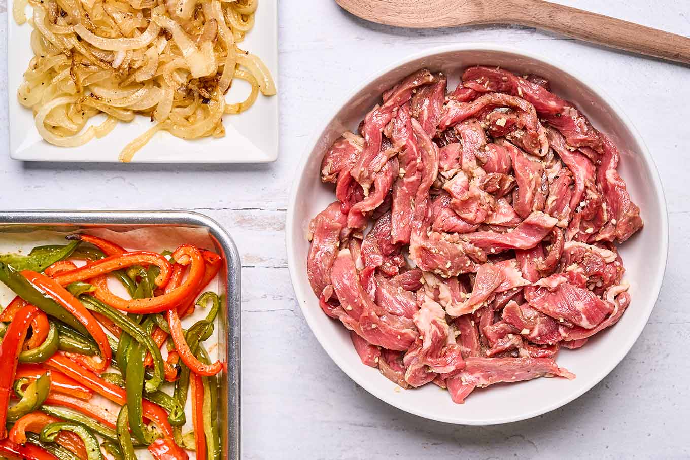 Thinly sliced steak in marinade alongside roasted peppers and sautéed onions for Philly cheesesteak sandwiches.