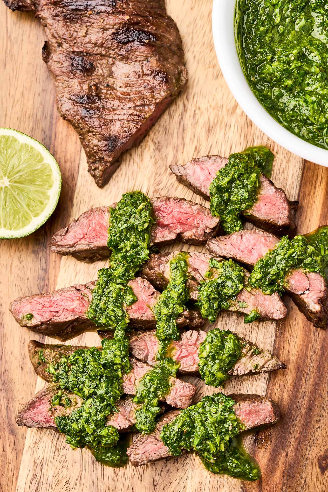 Sliced grilled steak with chimichurri sauce on a wooden board with a bowl of fresh chimichurri.
