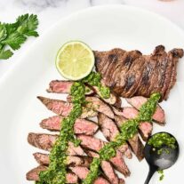 Grilled steak with chimichurri sauce sliced on a serving platter with a lime wedge and extra chimichurri.