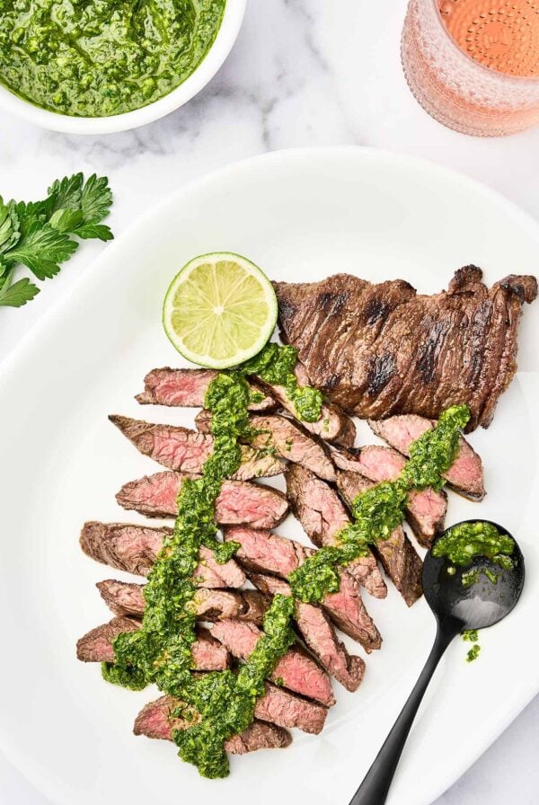Grilled steak with chimichurri sauce sliced on a serving platter with a lime wedge and extra chimichurri.