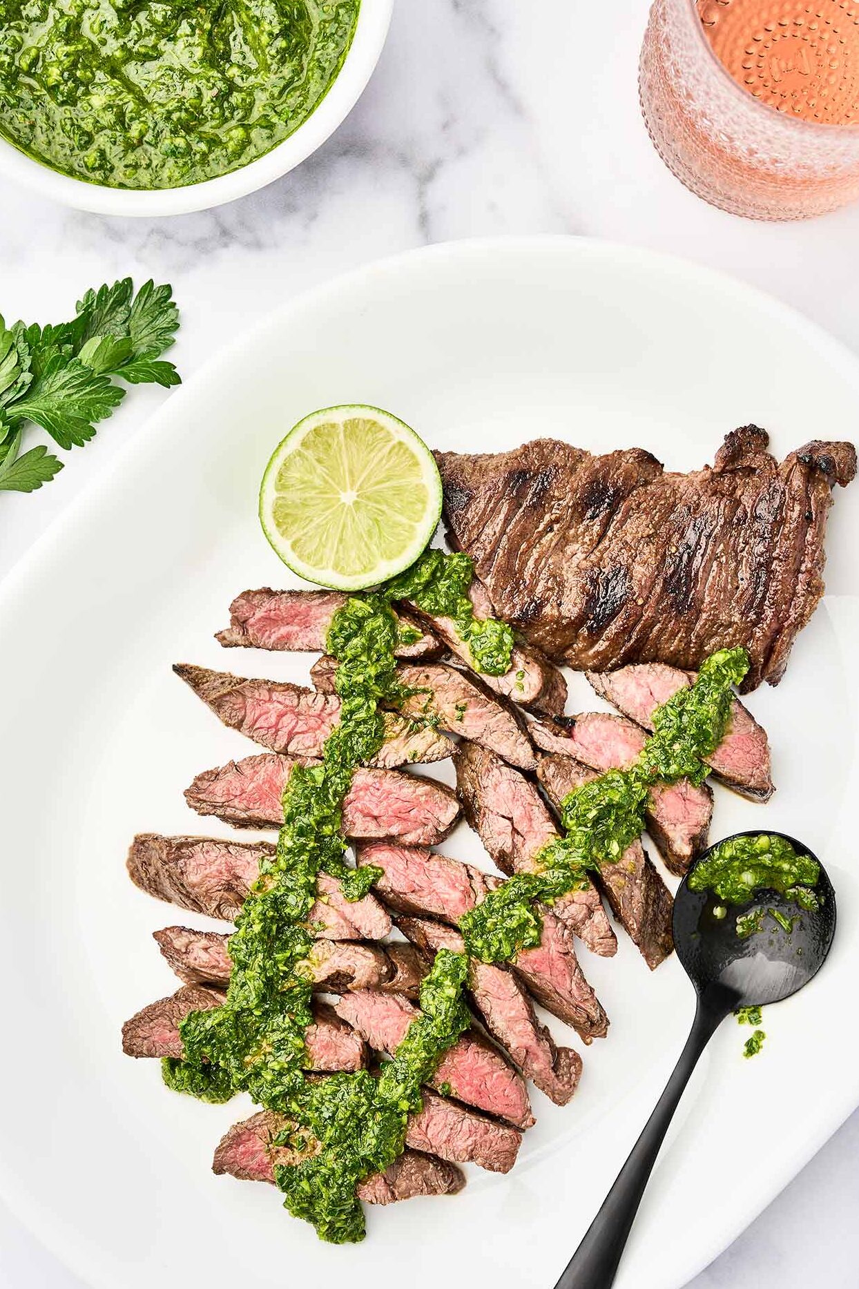 Grilled steak with chimichurri sauce sliced on a serving platter with a lime wedge and extra chimichurri.