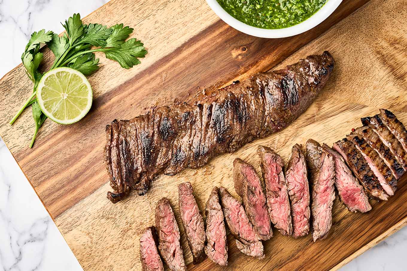 Grilled steak with chimichurri sauce with a whole skirt steak, sliced steak, and fresh chimichurri on a wooden cutting board.