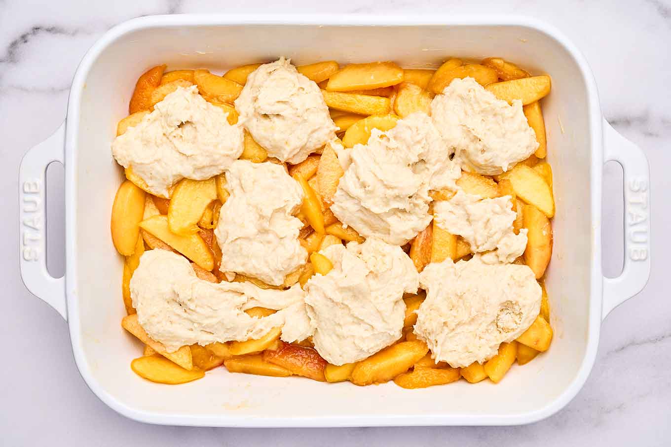 Spoonfuls of biscuit dough arranged over sliced peaches before baking fresh peach cobbler.