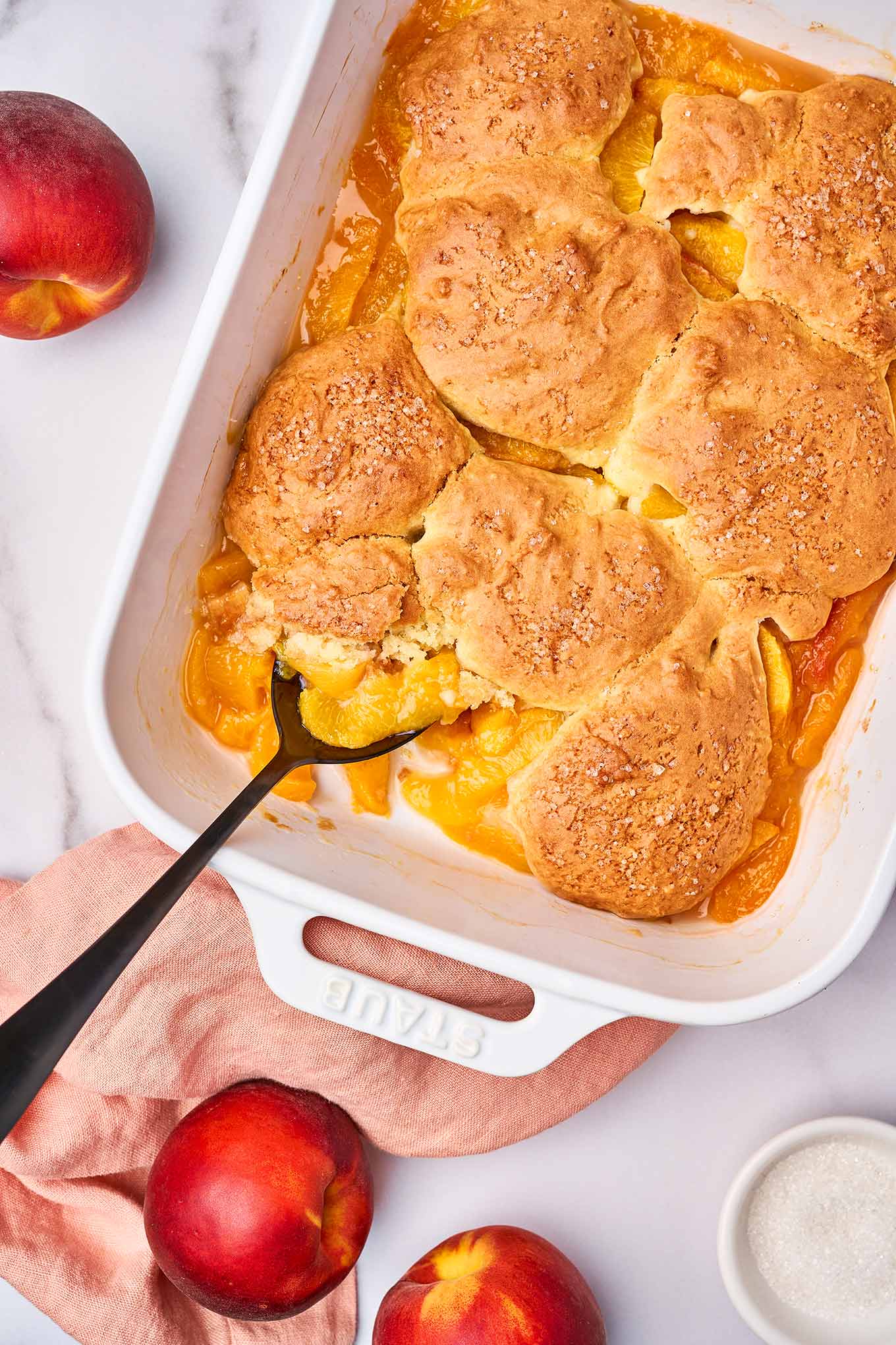Fresh peach cobbler with golden biscuit topping in a white baking dish, with one serving being spooned out.