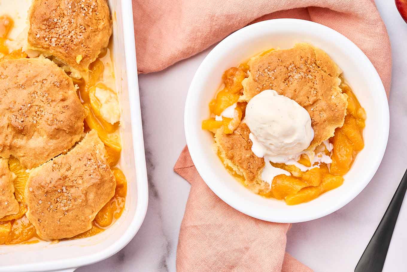 Fresh peach cobbler with biscuit topping and vanilla ice cream, served beside the baking dish.