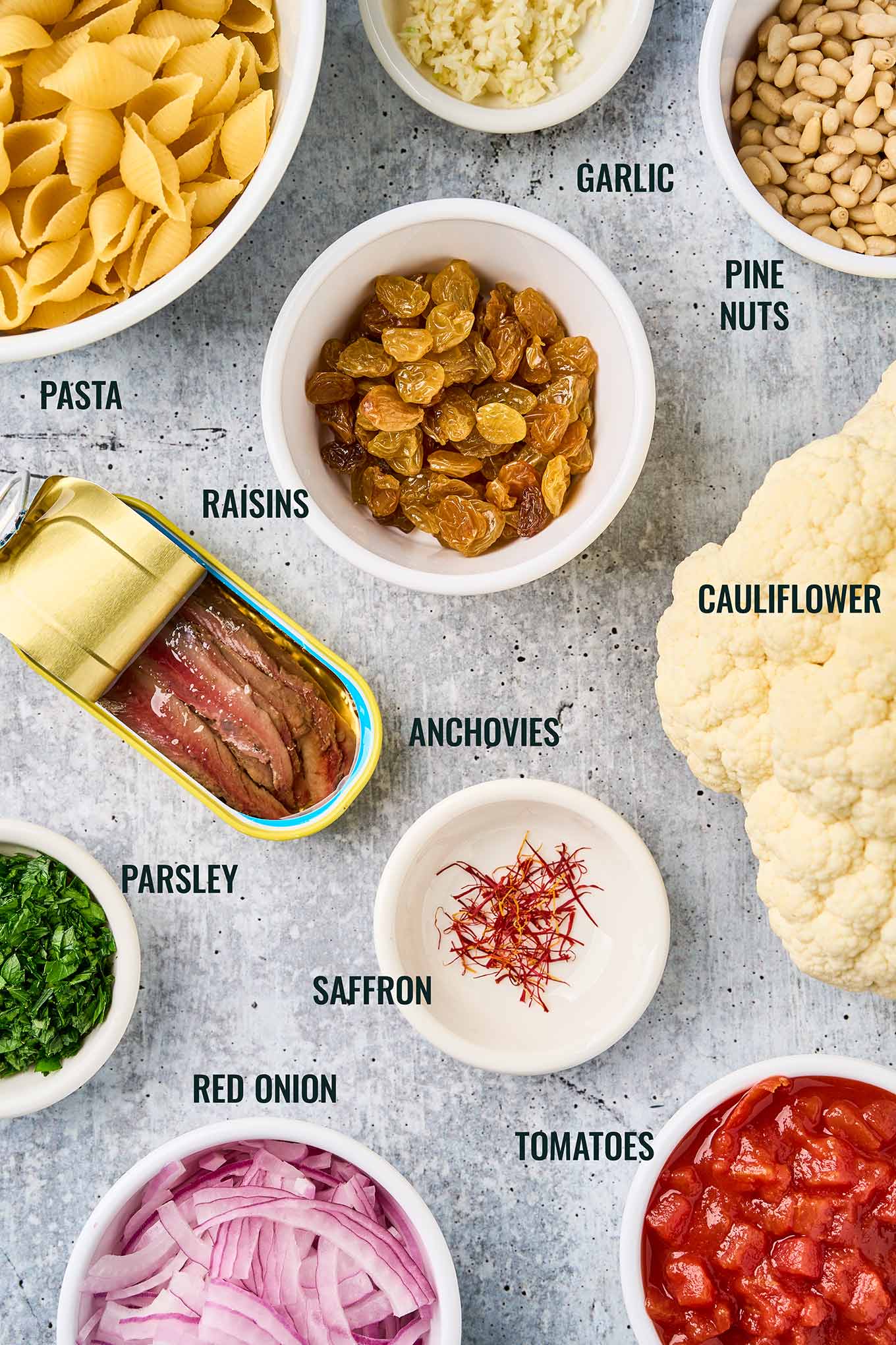 Ingredients for Sicilian pasta with cauliflower, including shell pasta, cauliflower, anchovies, saffron, raisins, pine nuts, tomatoes, onion, garlic, and parsley.