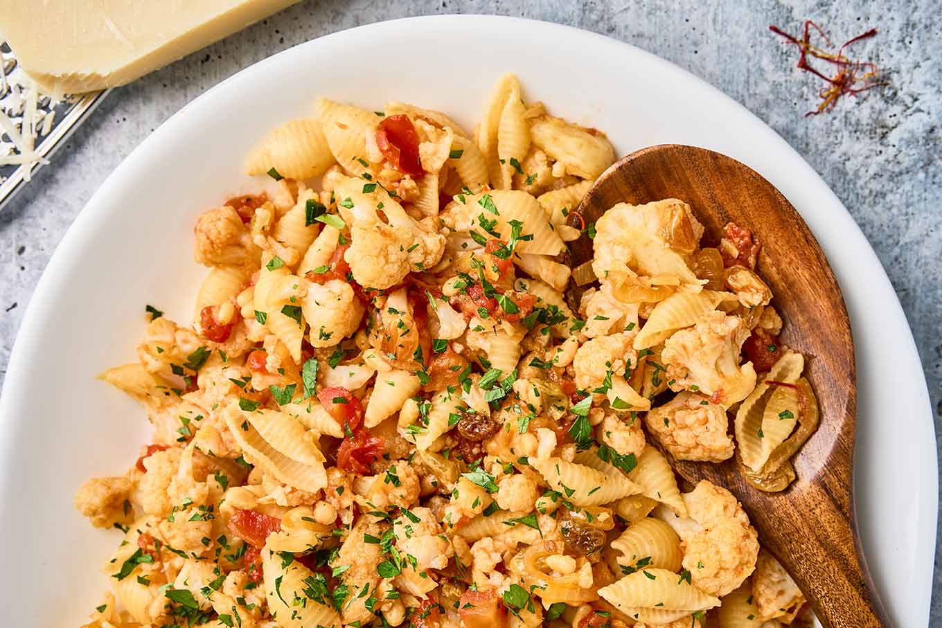 Close-up of Sicilian pasta with cauliflower, shell pasta, tomatoes, raisins, pine nuts, and parsley with a wooden serving spoon.