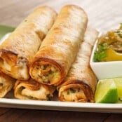 Baked chicken flautas with melted cheese, served on a white plate with lime wedges and a side of chunky tomatillo sauce garnished with fresh herbs.
