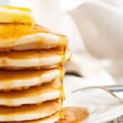 A side view of a stack of fluffy pancakes on a white plate, topped with a thick pat of butter and a drizzle of maple syrup.