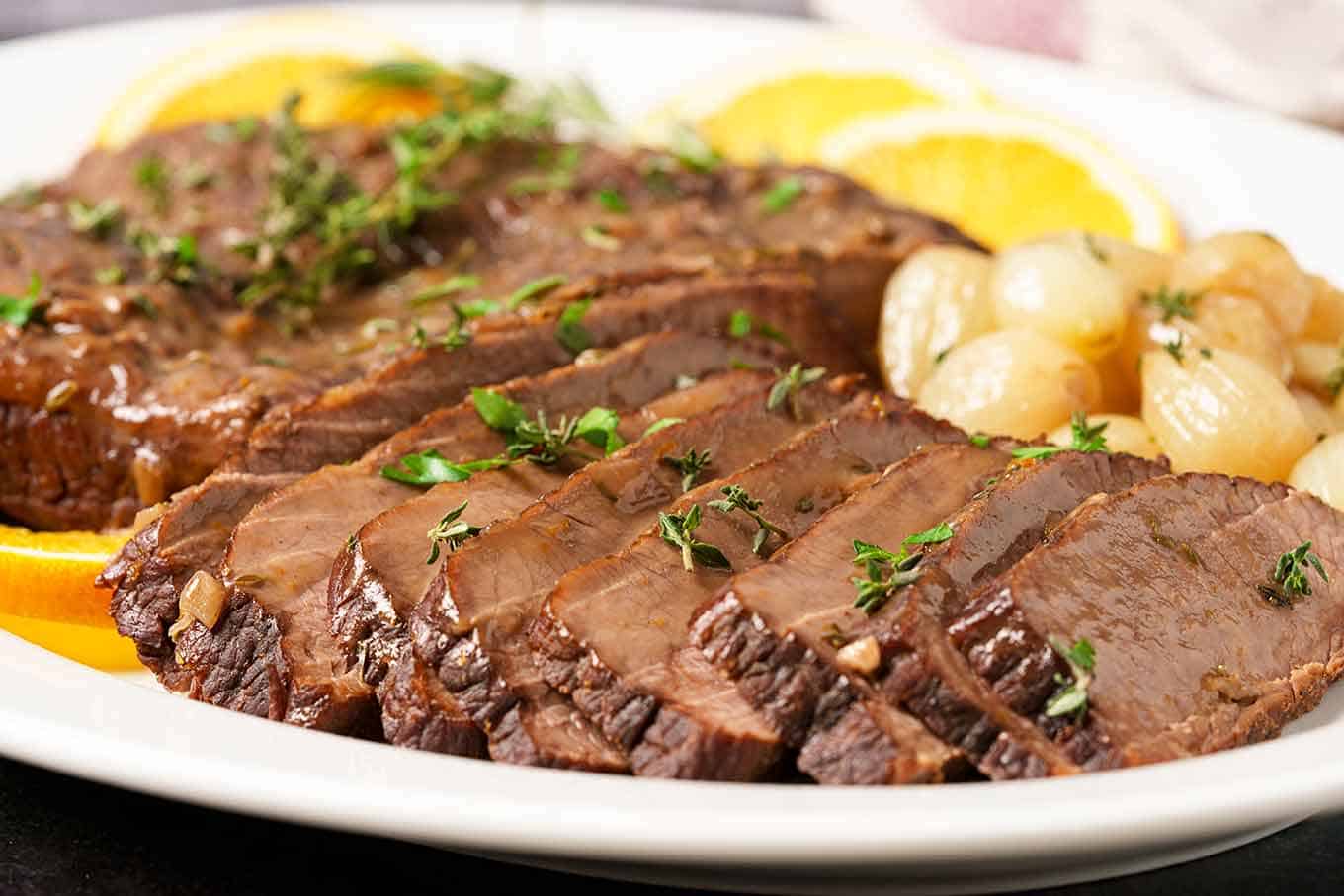 A close up of sliced beef brisket on a serving platter with pearl onions, parsley sprinkled on top for garnish.