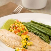 A plate of broiled catfish filet topped with fresh corn salsa, steamed green beans, and a lime wedge.