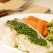 A close view of a broiled halibut fillet topped with kale pesto on a dinner plate with steamed carrots and a serving of pasta; a fork atop a dinner napkin to the rear.