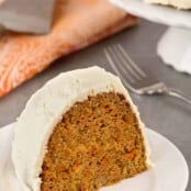 A slice of moist, spiced carrot cake with cream cheese frosting on a plate, the remainder of a cake on a stand in the background along with a fork and printed cloth napkin.