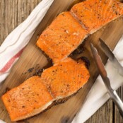 Two grilled salmon fillets on a cedar plank on top of a kitchen towel, metal tongs to the side.