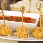 A close view of crispy chicken bites skewered with loop-ended bamboo skewers surrounding a small bowl of marinara dipping sauce.