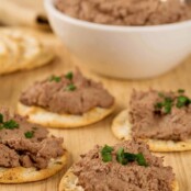Four water crackers topped with chicken liver paté and a sprinkling of chives on a wooden board, more paté and plain crackers in the background.