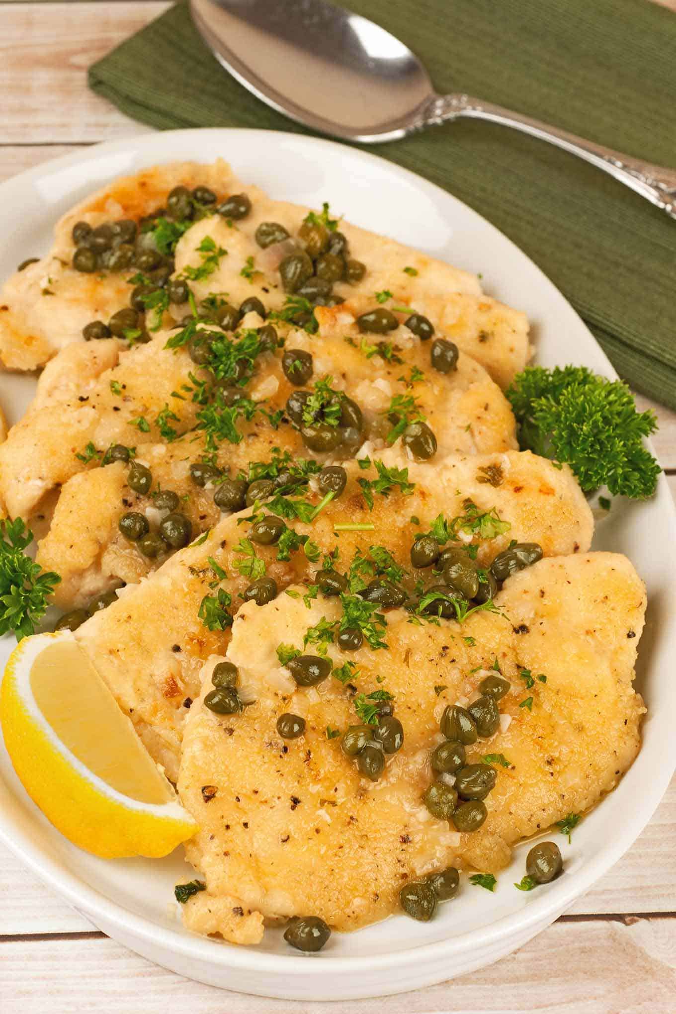 Chicken piccata on a serving plate garnished with capers, parsley, and a lemon wedge.