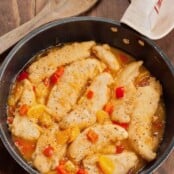 Golden brown chicken tenders simmered in a fresh orange pan sauce with chunks of orange and red bell pepper, served in a black skillet on a wooden surface.