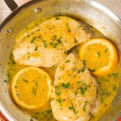 An overhead view of a stainless frying pan with two boneless chicken breasts, two orange slices, and orange pan sauce, sprinkled with chopped fresh tarragon.