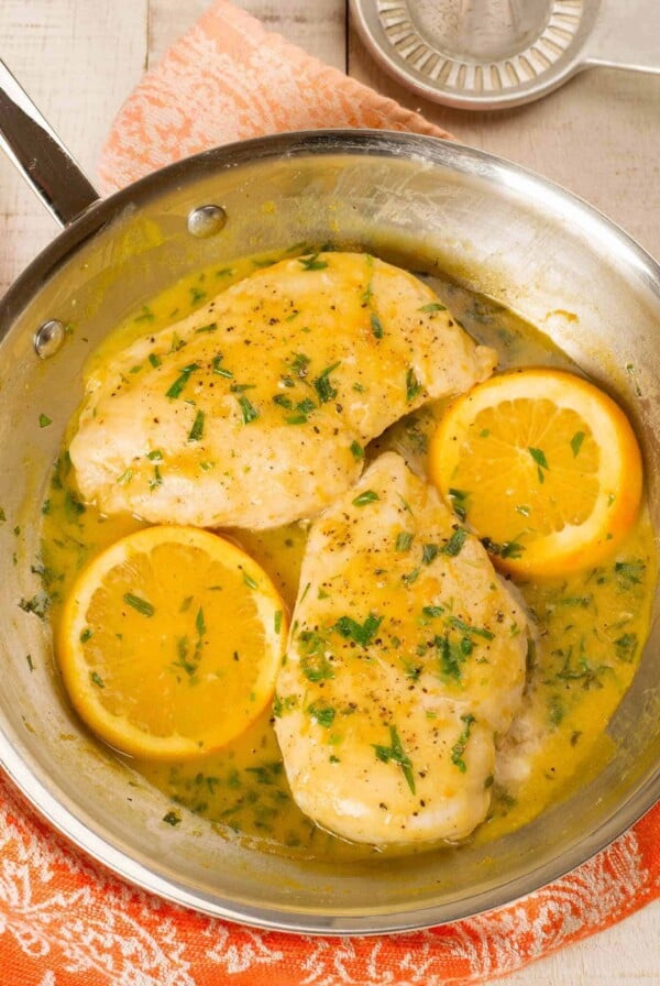 An overhead view of a stainless frying pan with two boneless chicken breasts, two orange slices, and orange pan sauce, sprinkled with chopped fresh tarragon.