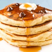 A stack of four chocolate chip pancakes topped with a few extra chocolate chips, a pat of butter, and a drizzle of maple syrup.