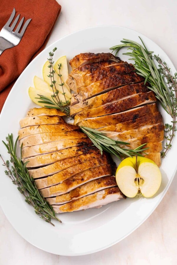 An overhead view of a carved turkey breast with golden brown skin on an oval serving platter garnished with fresh herbs and apple.