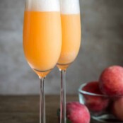 Two Bellini cocktails in Champagne flutes, foam on the top from the prosecco, a bowl of fresh peaches in the background.