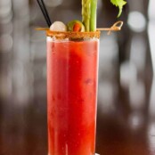 Tall glass of Bloody Mary cocktail garnished with celery stalk, pickled asparagus, green olive, and cocktail onion, served with a spiced rim and black straw.