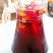 A clear glass pitcher filled with classic Spanish style sangria with ice, chunks of fruit, and a stirrer.