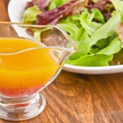 A glass gravy boat filled with vinaigrette dressing in front of a plate of fresh mixed greens on a wooden table.