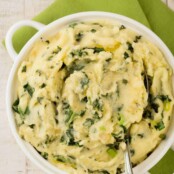 An overhead view of a white casserole filled with creamy mashed potatoes mixed with sautéed kale, sweet onion, and scallions (colcannon), and a serving spoon.