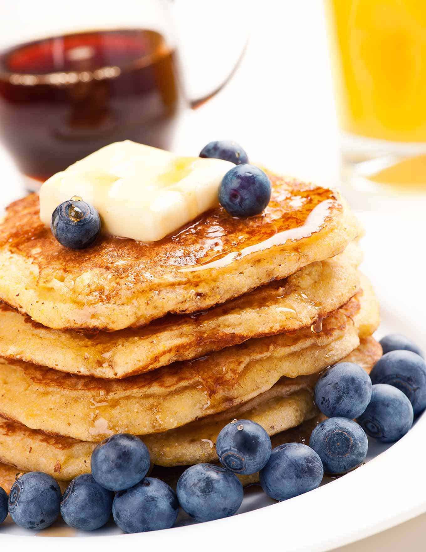 A stack of cornmeal pancakes on a plate topped with a pat of butter, maple syrup and fresh blueberries.