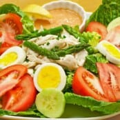 A serving of crab Louie salad, lettuce, tomatoes, hard cooked eggs, crab, cucumber, asparagus, dressing, and a lemon wedge