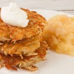 Golden crispy potato latkes stacked on a white plate, topped with sour cream and served with applesauce.