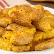 A stack of golden, crispy smashed potatoes on a white plate, seasoned with salt and pepper.