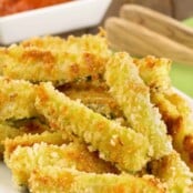 Crispy oven-fried zucchini sticks coated in panko crumbs and grated Parmesan stacked on a serving plate with marinara dipping sauce in the background.