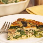 A close view of a slice of crustless morel mushroom and ramp quiche of a plate, a fork to the side and the dish with more quiche in the background.