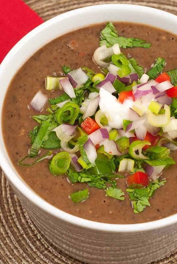 A close-up photo of a cup of black bean soup topped with diced fresh vegetables including jalapeno and red bell peppers, red onion, scallions, and cilantro on a rattan placemat.