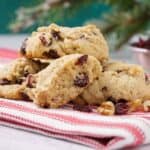Cranberry walnut cookies on a folded red and white towel with dried cranberries and nuts scattered in front, a dish of dried cranberries in the background.