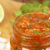 An open jar of homemade salsa, garnished with chopped cilantro, out of focus ingredients on a cutting board in the background.