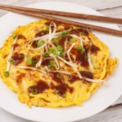 Golden Egg Foo Young omelet topped with brown gravy, bean sprouts, and sliced green onions on a white plate with chopsticks.