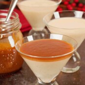 Three parfait glasses with eggnog panna cotta, the first one topped with homemade caramel rum sauce, a jar of sauce to the left, Christmas table decor in the background.