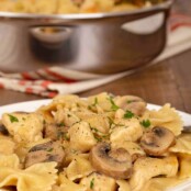 A serving plate of farfalle combined with mushrooms and bite-sized pieces of chicken breasts in a creamy marsala sauce, a sauté pan with more in the background.