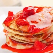 Stack of fluffy ricotta pancakes topped with vibrant strawberry-orange sauce and chunks of fresh strawberries, served on a white plate.