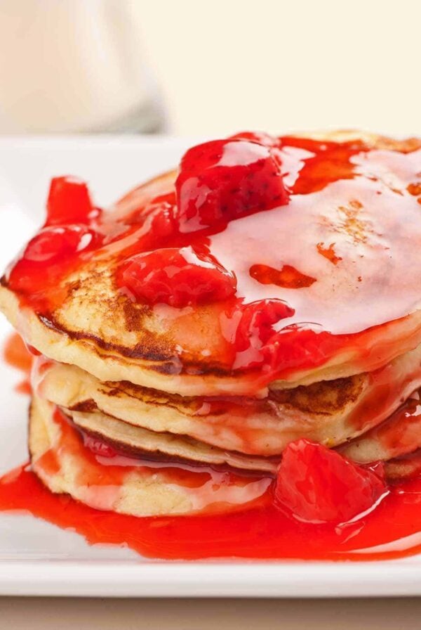 Stack of fluffy ricotta pancakes topped with vibrant strawberry-orange sauce and chunks of fresh strawberries, served on a white plate.