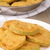 Crispy, cornmeal crusted, fried green tomatoes on a serving plate, a pan with more in the background.
