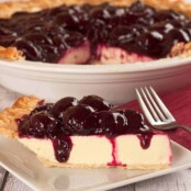 A dessert plate with a slice of Greek yogurt pie topped with fresh cherry topping, the remainder of the pie in a white pie plate in the background.