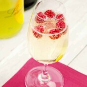 A champagne flute filled with a sparkling cocktail of prosecco and limoncello with fresh raspberries floating on top on a pink cocktail napkin, bottles in the background.