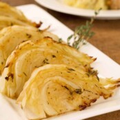 Wedges of herb and garlic roasted cabbage on a serving plate, garnished with thyme, dinner plate in the background.