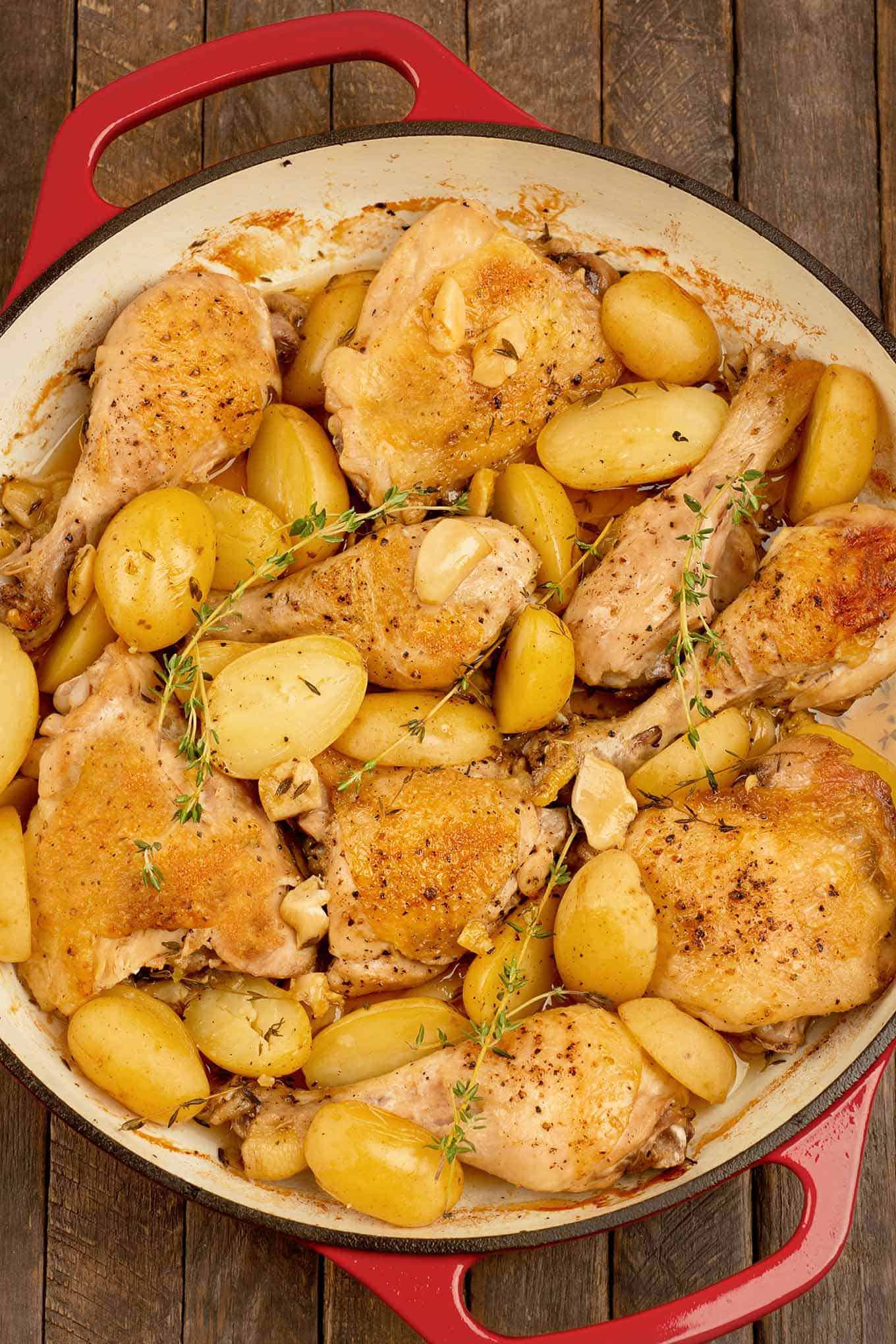 A cast iron skillet filled with roasted chicken thighs and drumsticks, potatoes, garlic, and sprigs of fresh thyme.